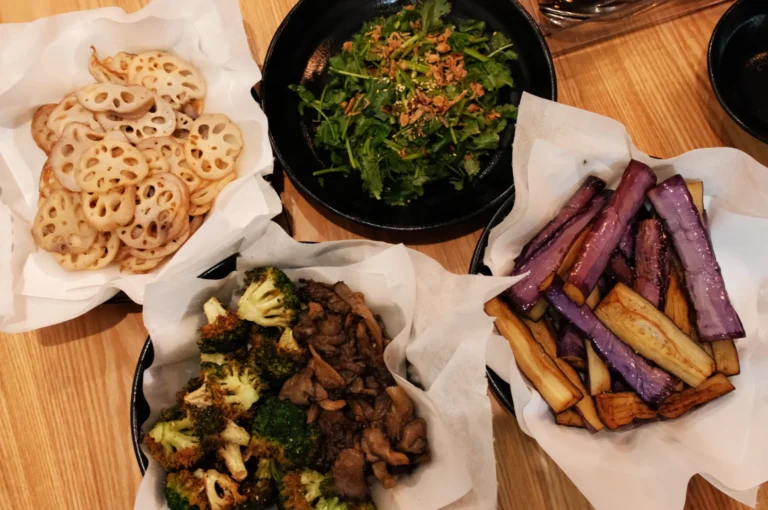 A variety of dishes including sliced lotus root, a salad with greens and sesame, sautéed broccoli with meat, and roasted eggplant.