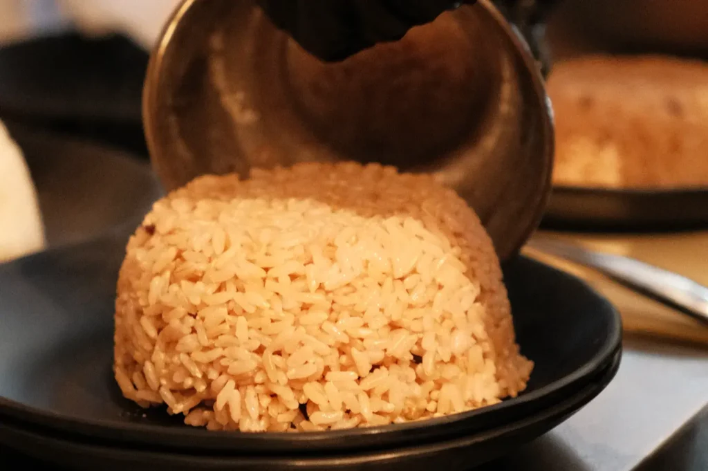 A close-up of a person pouring cooked rice from a metal bowl onto a black plate, forming a mound of rice.