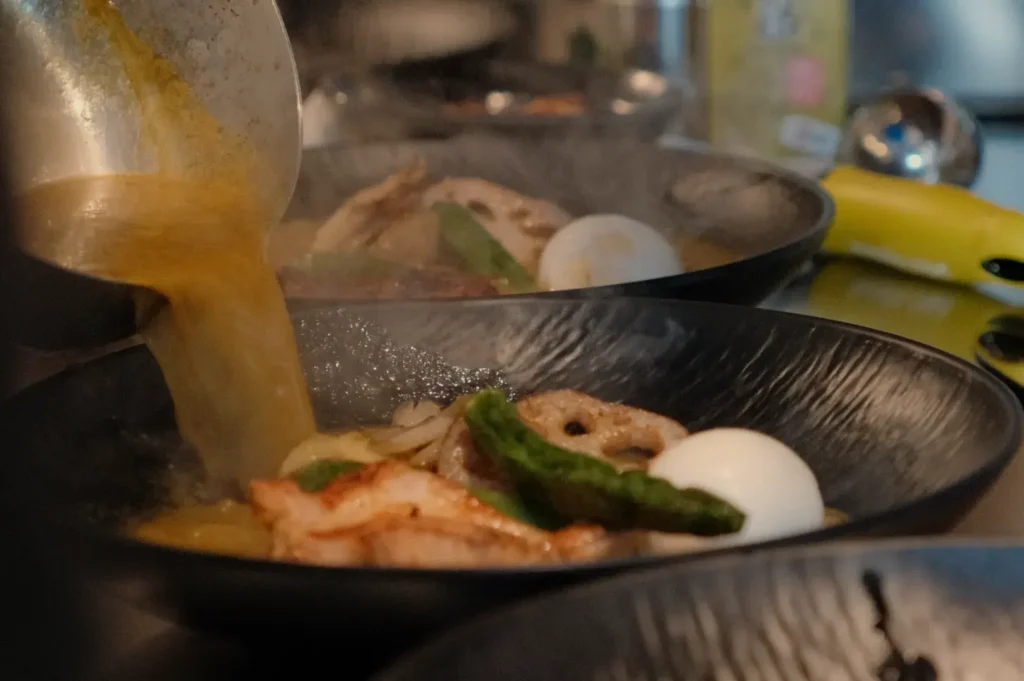 A bowl of soup curry being prepared, with broth being poured over noodles, vegetables, and an egg.