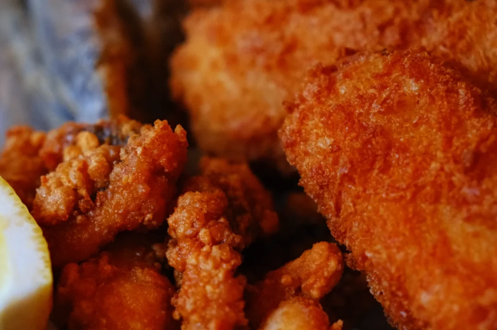 A close-up image of crispy fried chicken pieces, including smaller pieces and a larger fillet, with a slice of lemon in the corner.