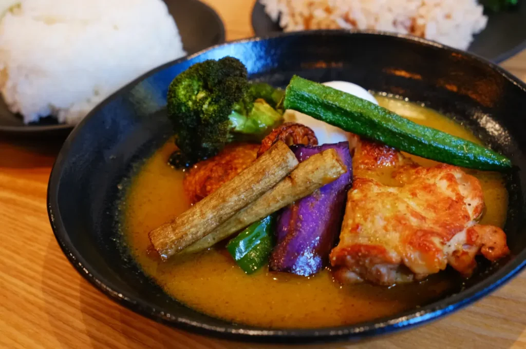 A bowl of soup curry featuring chicken, vegetables like broccoli and okra, and a side of rice.