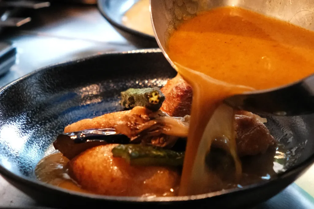 A dish being served with a rich, golden sauce being poured over it, featuring various ingredients such as vegetables and possibly meat, in a black bowl.