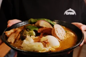 A bowl of soup curry topped with various vegetables, a soft-boiled egg, and slices of meat, held by a person wearing a black shirt.