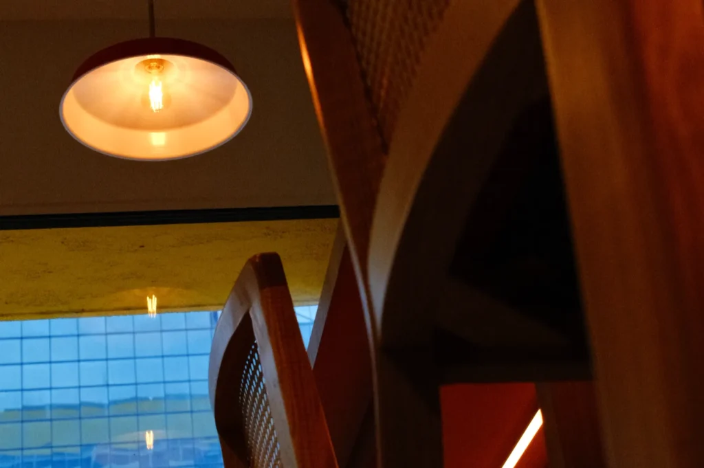 A close-up view of wooden chairs with a hanging light above. The background features a window partially obscured by the chairs, showing a grid pattern with blue and yellow tones.