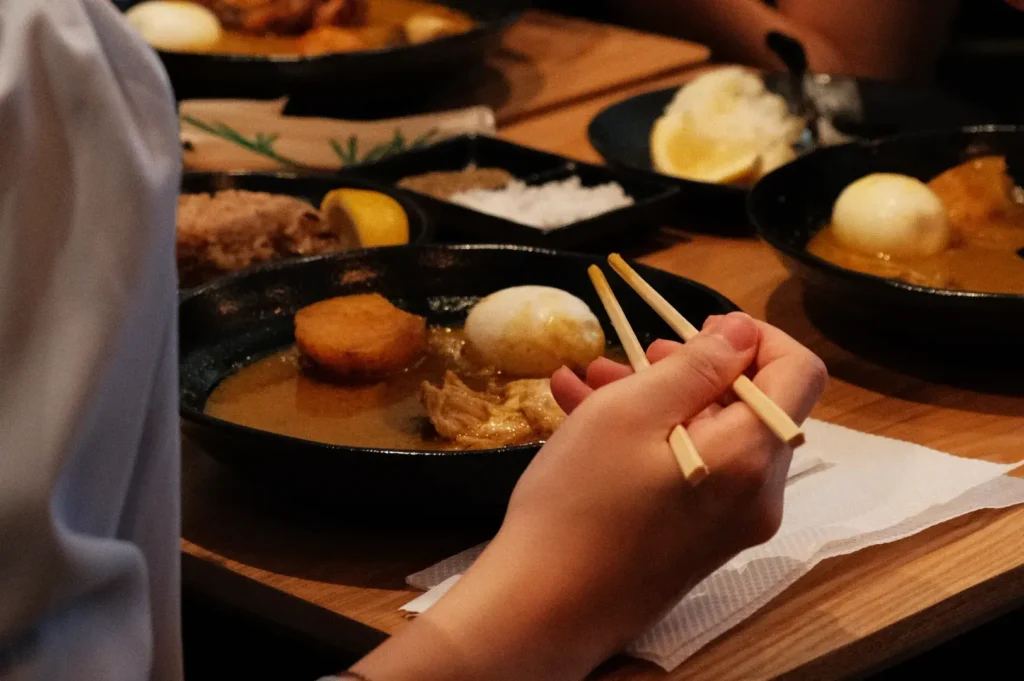 A hand holding chopsticks over a bowl of Japanese curry with various ingredients, including an egg and vegetables, on a wooden table.