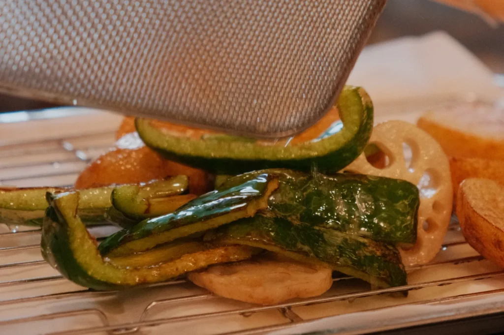 A variety of sliced and flash fried vegetables, including zucchini and lotus root, resting on a rack with a metal strainer above them.