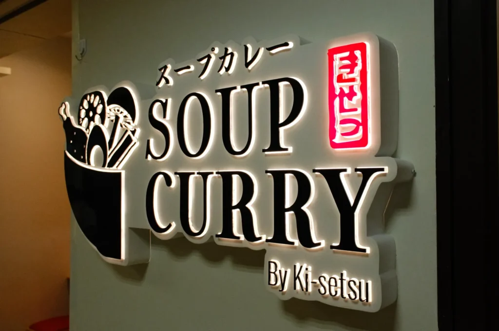 A sign that reads "SOUP CURRY" with Japanese text above it, featuring a graphic of a bowl with food, illuminated in a modern font.