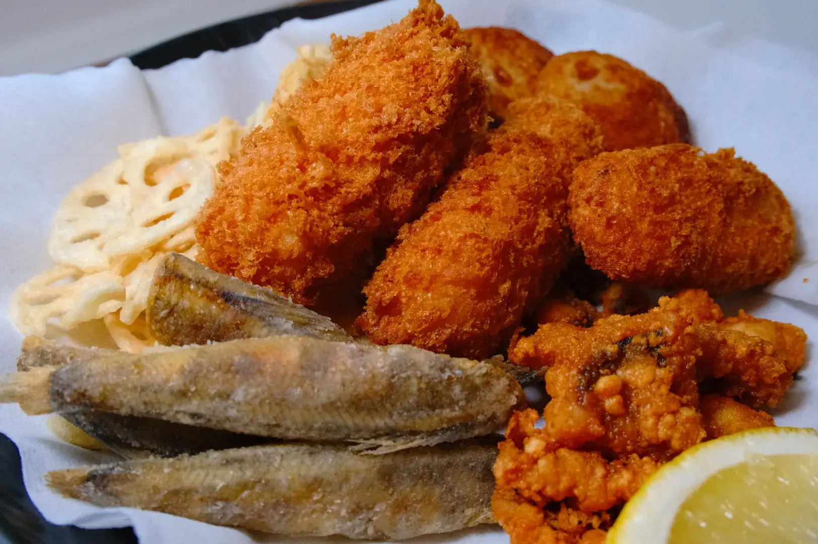 A plate of assorted fried foods, including crispy fish, croquettes, and lotus root slices, with a lemon wedge.