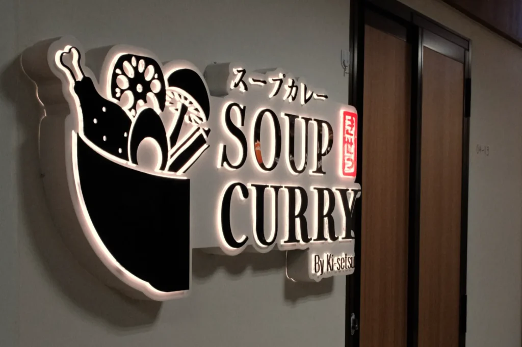 Signage for "SOUP CURRY" featuring illustrations of various ingredients, with Japanese text and the name "By Katsu" underneath.