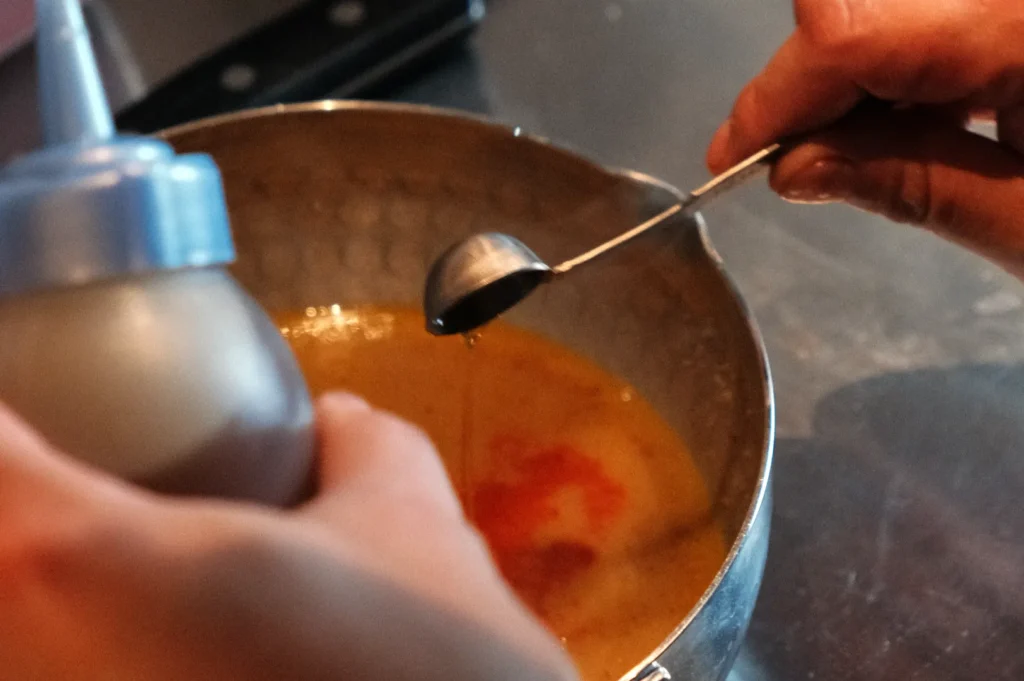 A person holding a squeeze bottle and a measuring spoon over a metal bowl containing a yellow sauce.
