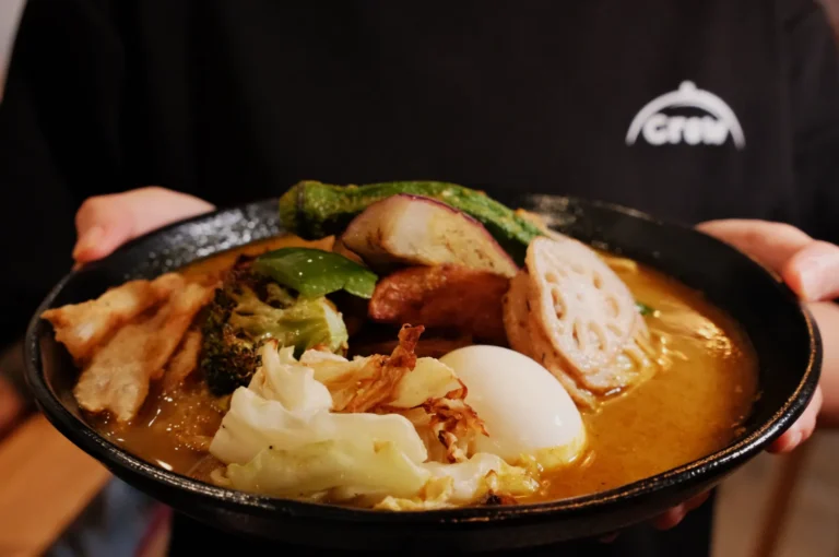 A bowl of soup curry with chicken, broccoli, green beans, eggplant, and okra, served with rice in the background.