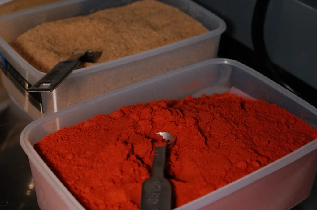 Containers of spices, one with reddish powder and the other with brown granules, both with measuring spoons placed inside.