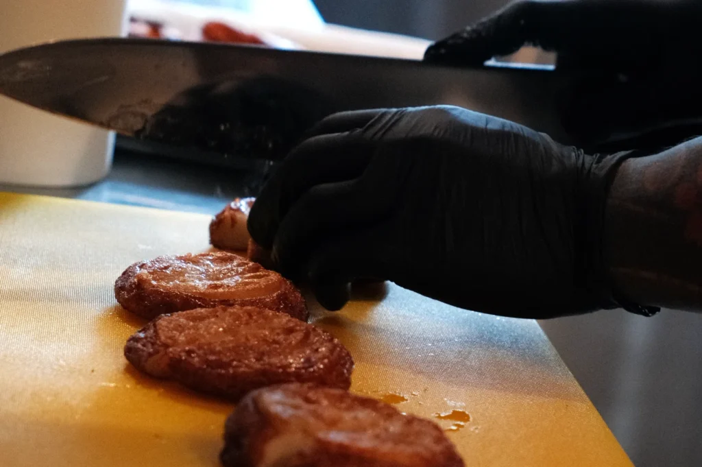 A person's hand in a black glove is slicing circular, fried food items on a yellow cutting board.