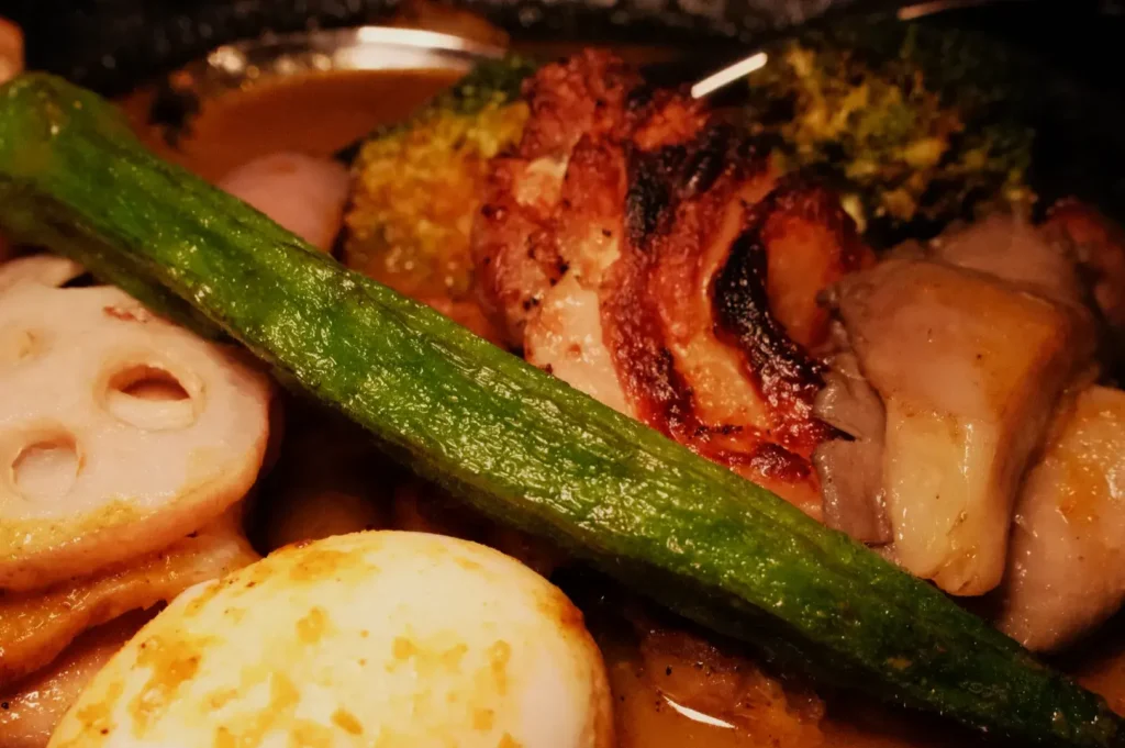 A close-up view of soup curry featuring grilled meat, broccoli, sliced lotus root, and okra, all served in a savory sauce.