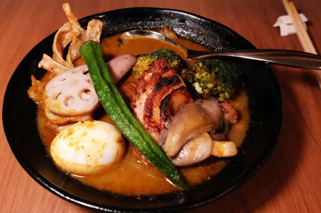 A bowl of soup curry with various toppings including sliced meat, vegetables, an egg, and a spoon.