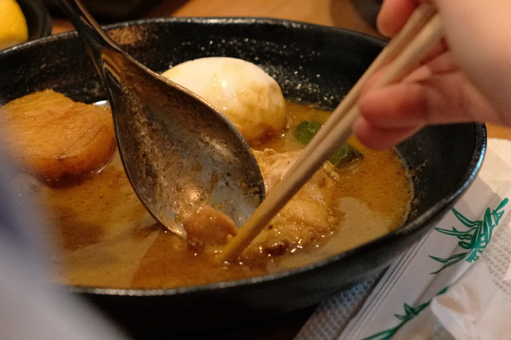 A person using chopsticks to eat from a bowl of ramen, with a soft-boiled egg and slices of meat visible in the broth.