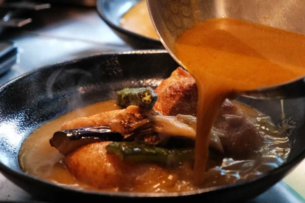 A pot of food being served, with a rich sauce being poured over vegetables and a protein in a skillet.