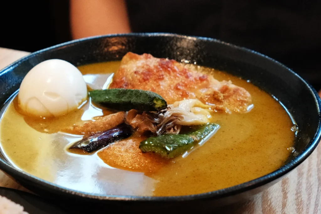 A bowl of soup curry with a boiled egg, pieces of chicken, and assorted vegetables including green beans and eggplant.