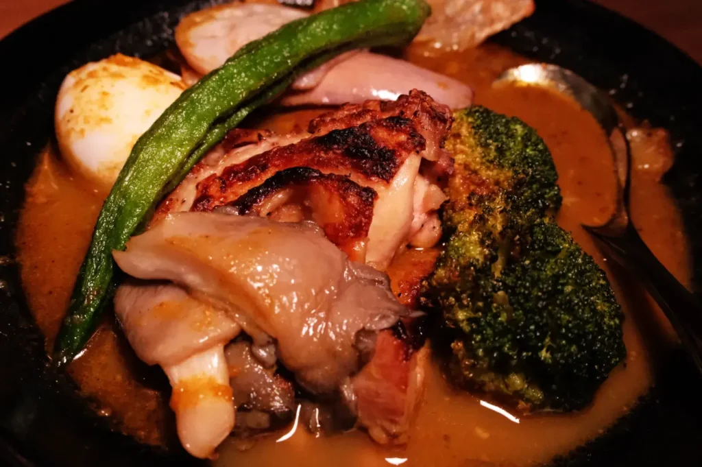 A plate of soup curry featuring grilled chicken, broccoli, a green pepper, and an egg in a savory sauce.