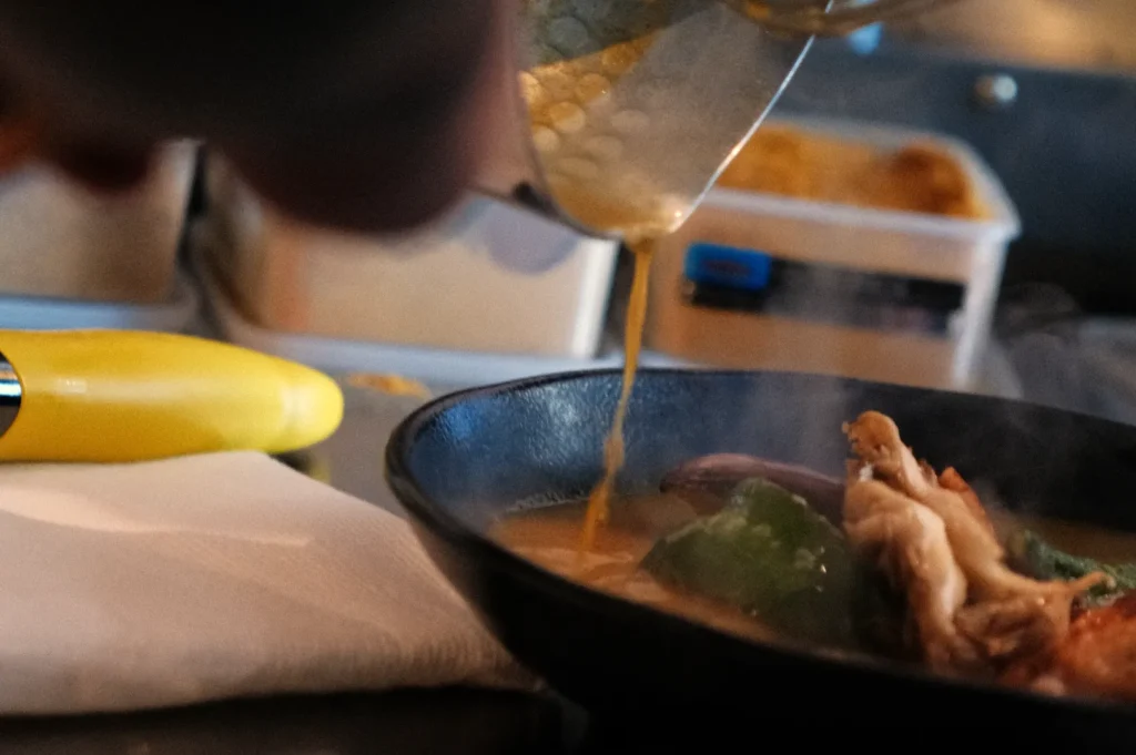 A person pouring liquid into a bowl containing food, with some vegetables and meat visible.