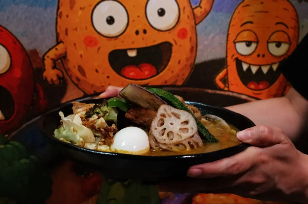 A person holding a bowl of soup curry with various toppings, including a slice of lotus root, vegetables, and an egg, in front of a colorful cartoon background.