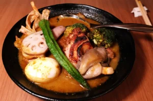 A bowl of ramen with various toppings, including grilled chicken, broccoli, lotus root, okra, and a boiled egg in a rich, brown broth.