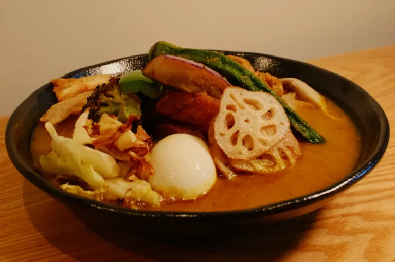 A bowl of soup curry topped with various vegetables, including lotus root, broccoli, and okra, alongside a soft-boiled egg.