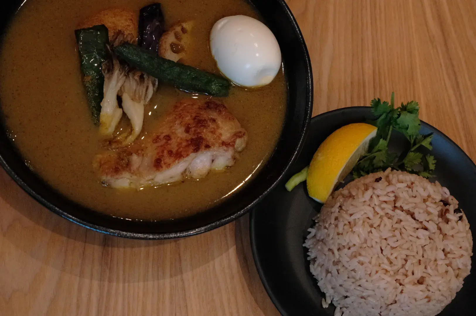 A bowl of curry with chicken, vegetables, and a soft-boiled egg, accompanied by a plate of brown rice garnished with a lemon wedge and cilantro.