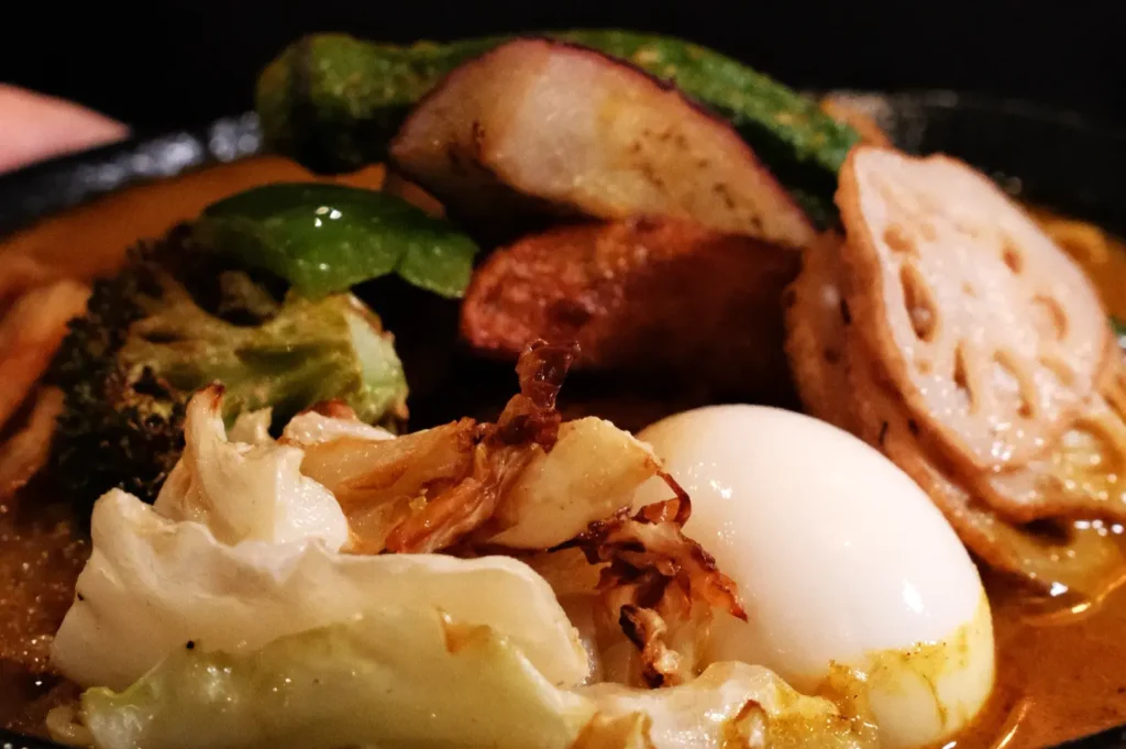 A close-up view of a plate of soup curry featuring various vegetables, including lotus root, broccoli, and leafy greens, alongside a soft-boiled egg in a flavorful sauce.