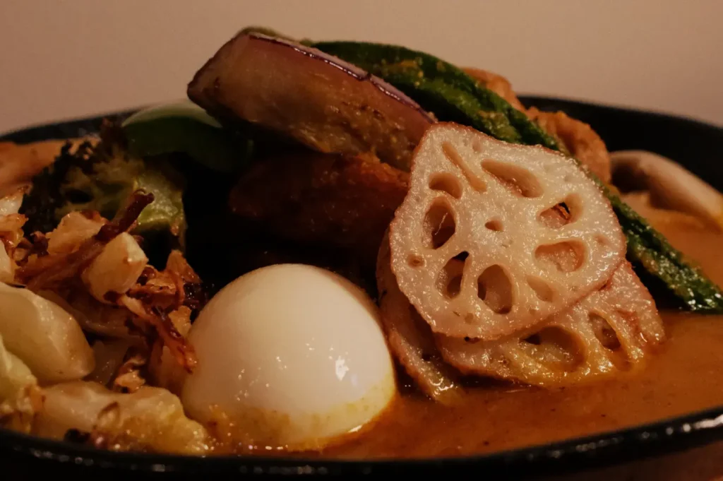 A plate of soup curry featuring various toppings including lotus root, a soft-boiled egg, fried vegetables, and a creamy broth.