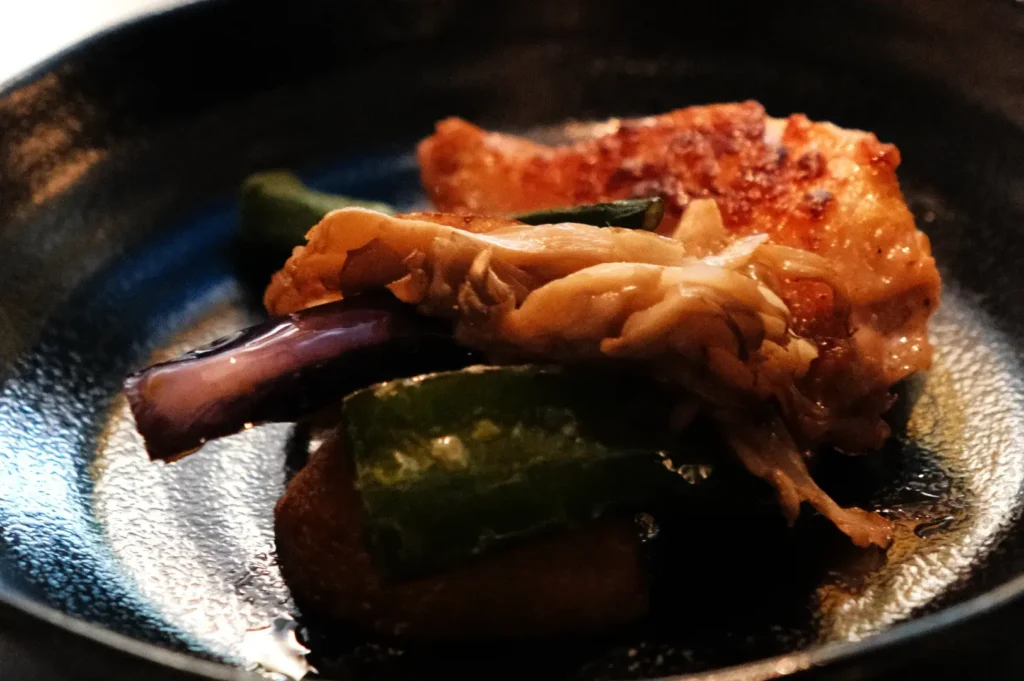A plate of food featuring a piece of chicken, eggplant, and green peppers arranged on a dark plate.