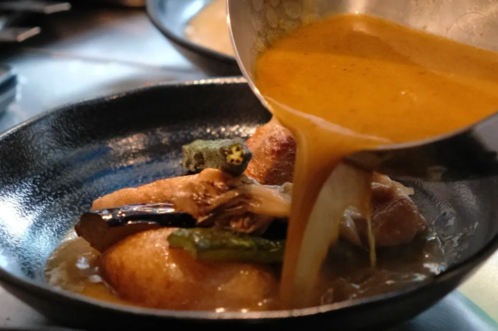 A close-up of a soup curry being served, with sauce being poured over vegetables and a type of bread or dumpling in a black bowl.