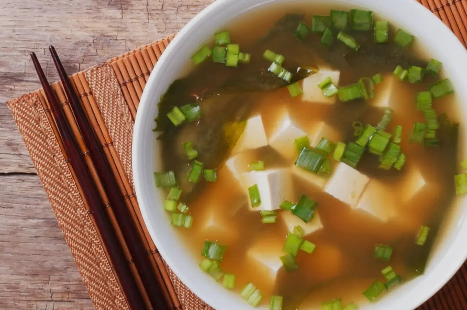 A bowl of soup with tofu and green onions, placed on a bamboo mat with chopsticks beside it.