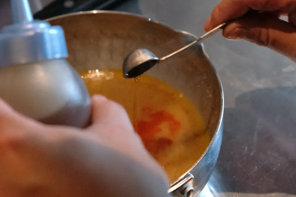 A person pouring a liquid from a bottle into a pot while holding a measuring spoon.