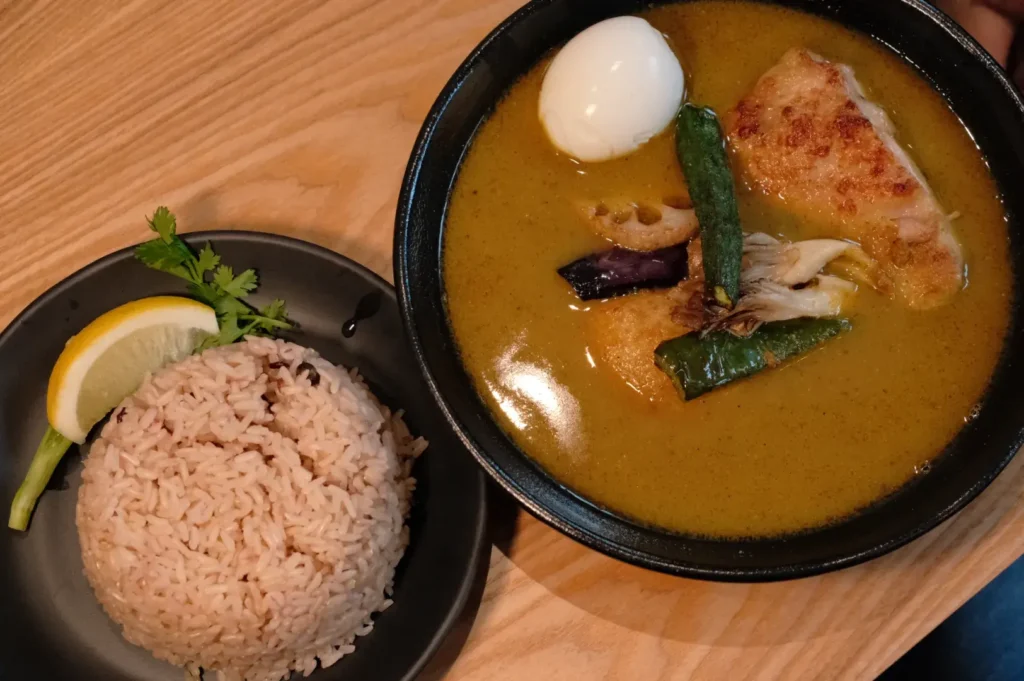 A bowl of curry soup with chicken, vegetables, and a soft-boiled egg, accompanied by a side of brown rice and a lemon wedge on a black plate.