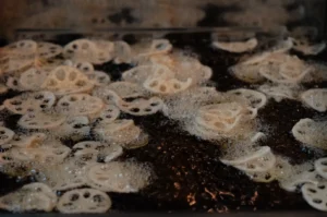 Thin lotus root slices with distinctive holes sizzle in bubbling oil in a dark pan, their pale surfaces crisping in a lively close-up of cooking.