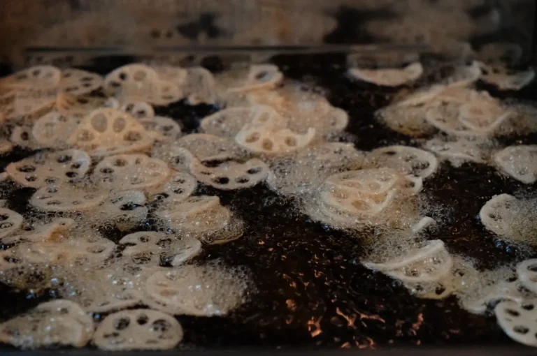 Thin lotus root slices with distinctive holes sizzle in bubbling oil in a dark pan, their pale surfaces crisping in a lively close-up of cooking.