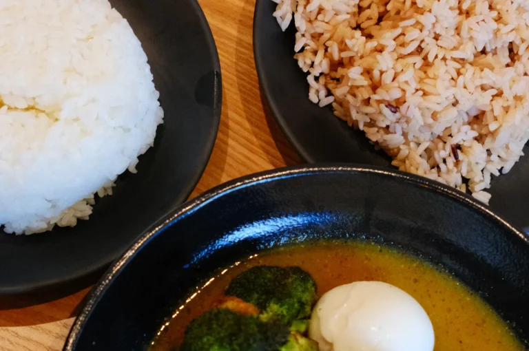 A plate of food featuring a black bowl of broccoli in yellow sauce with a soft-boiled egg, and two plates of rice, one white and one brown.