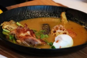 A bowl of rich broth with assorted ingredients, including pieces of meat, vegetables, and a poached egg.