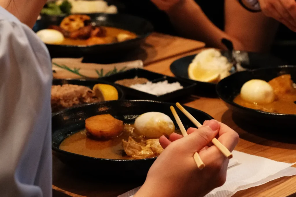 A close-up of hands holding chopsticks, with a bowl of ramen and various side dishes on a table.