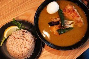 A bowl of curry with chicken, boiled egg, and vegetables, served alongside a plate of brown rice and a lemon wedge.
