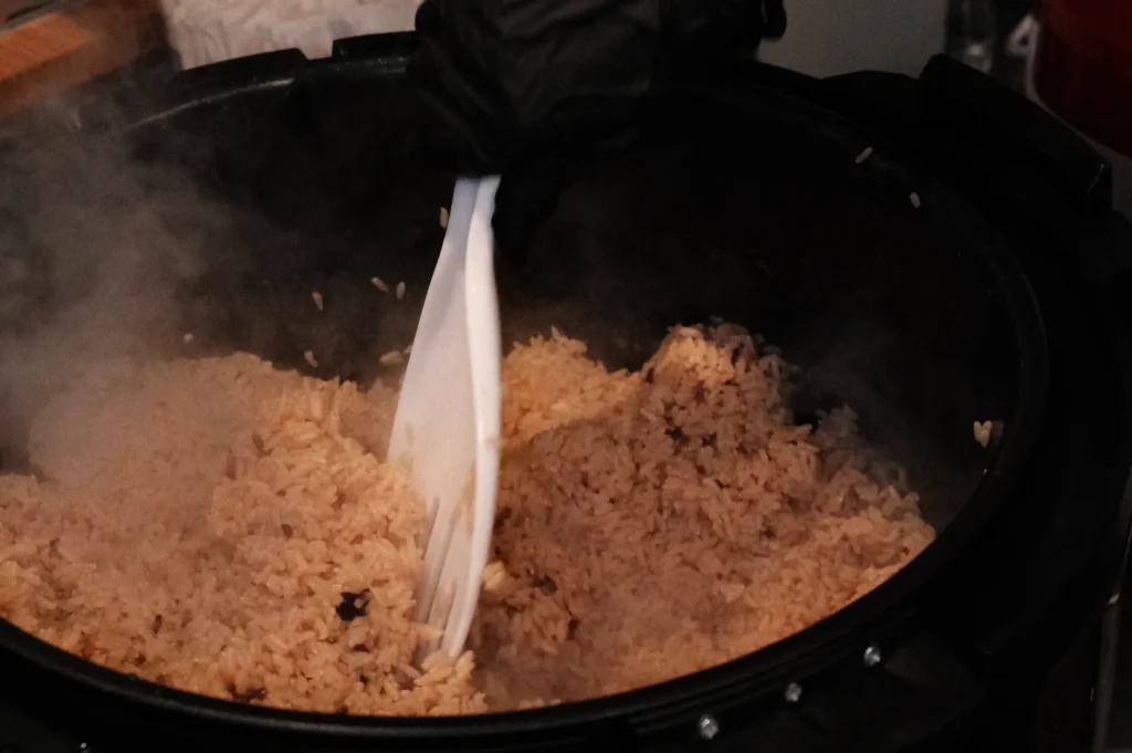 A close-up of a pot of rice being stirred with a white spatula, with steam rising from the pot.