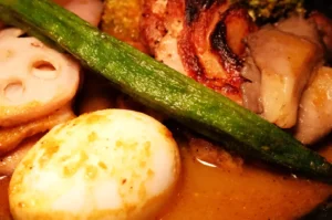 A close-up of a dish featuring cooked meats, an egg, okra, and sliced lotus root in a flavorful broth.