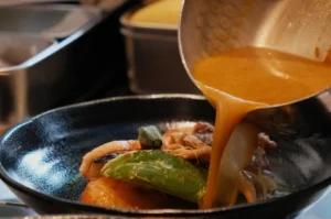 A dish being served with sauce being poured over it, featuring various ingredients like vegetables and meat in a bowl.