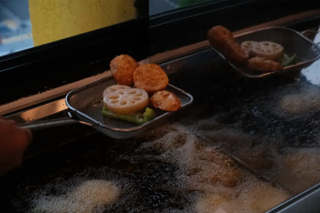 A cook lifts wire baskets of golden fried lotus root, patties, and other snacks from a sizzling griddle, creating a warm, busy street-food scene.