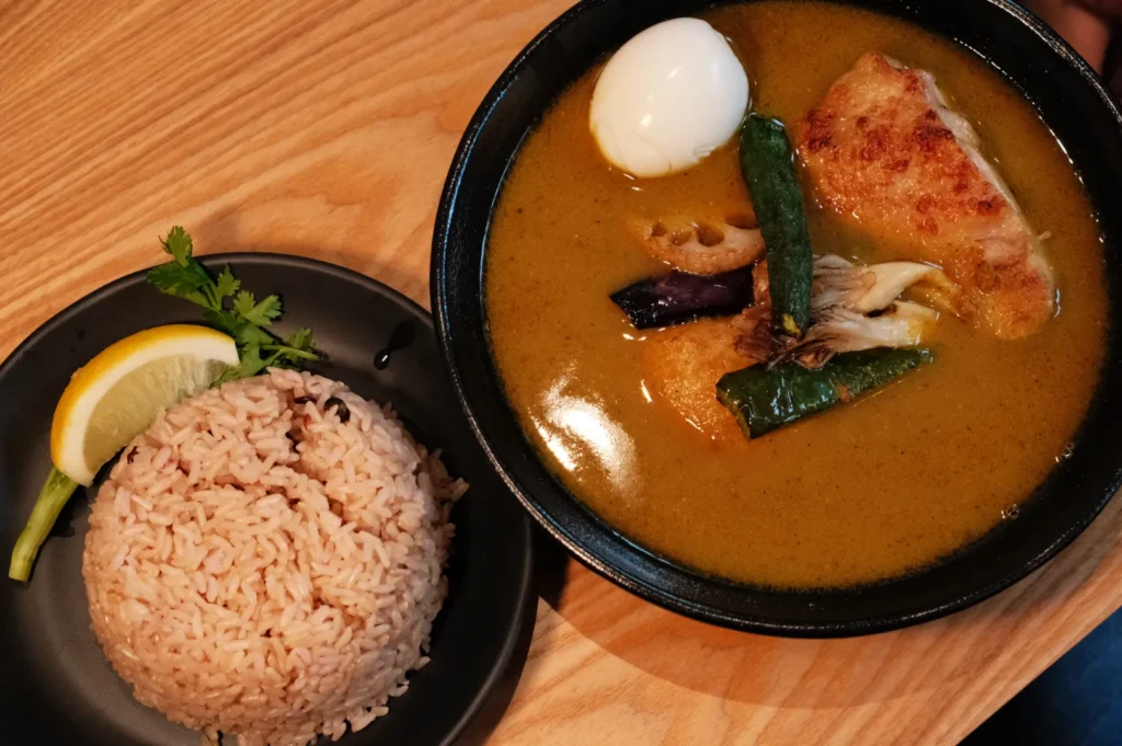A bowl of curry with vegetables and a fried egg, accompanied by a plate of brown rice and a lemon wedge.