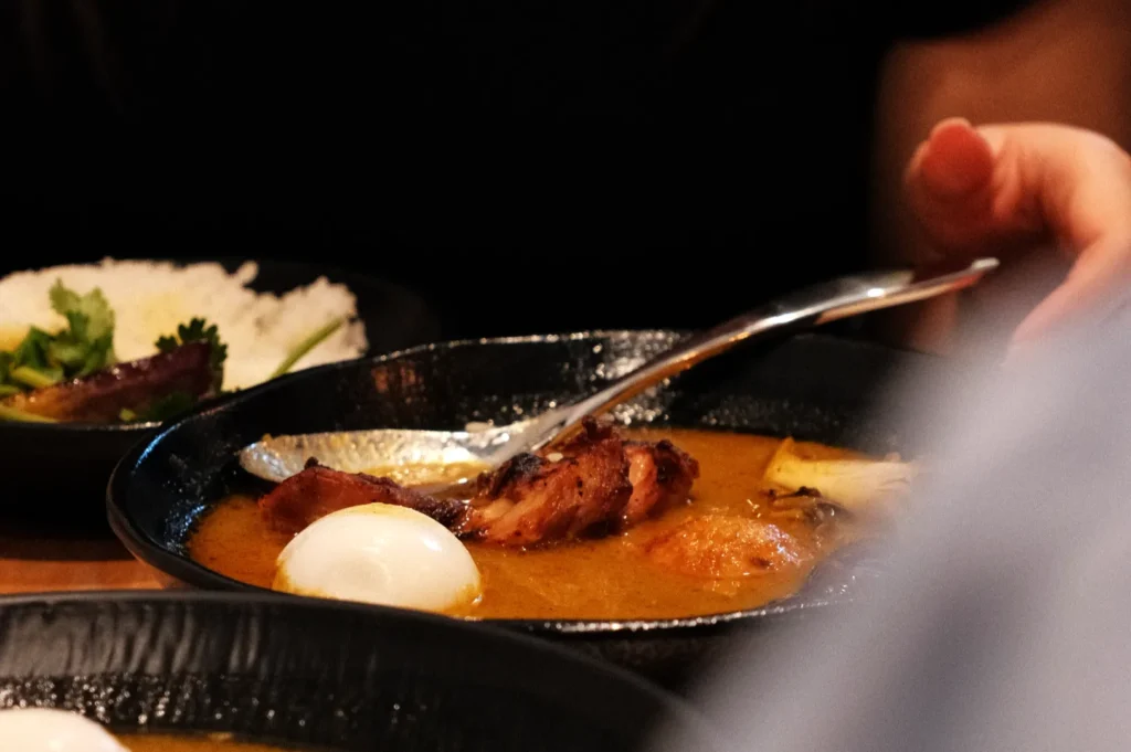 A bowl of rich curry with pieces of meat, boiled eggs, and a spoon, with a side of rice and greens in the background.