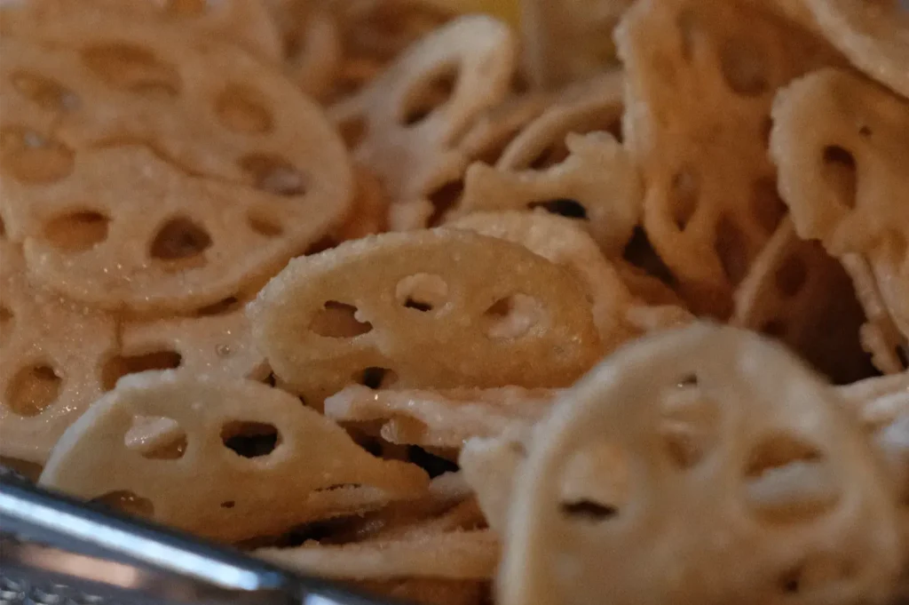 Close-up of golden-brown fried lotus root chips, their distinctive round slices showing delicate holes and crisp texture, piled in a metal bowl.