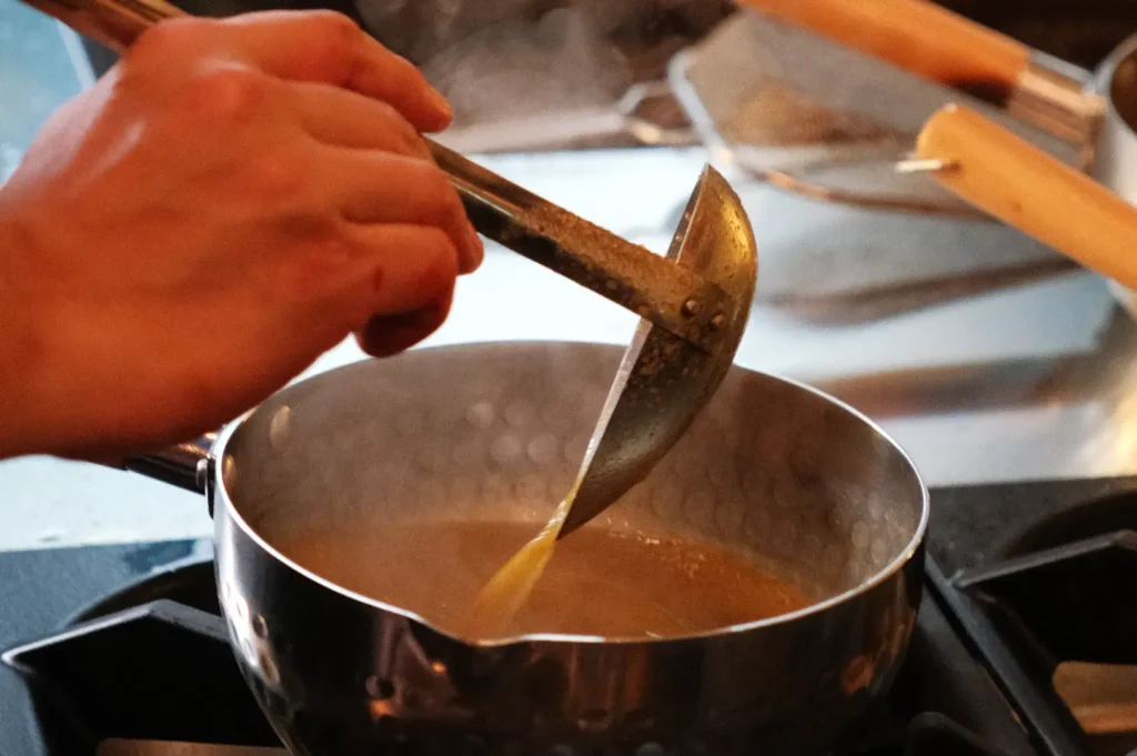 Close-up of hands using a metal ladle to pour steaming brown sauce into a stainless-steel saucepan on a stovetop, creating a warm cooking scene.