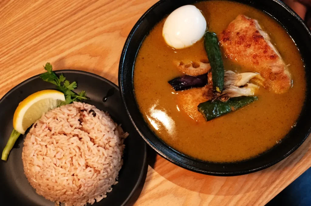 A bowl of curry with a piece of fried chicken, vegetables, and a soft-boiled egg, served with a side of brown rice and a lemon wedge.